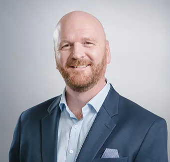 Smiling bald man with ginger beard wearing a navy blazer and blue shirt against light gray background.