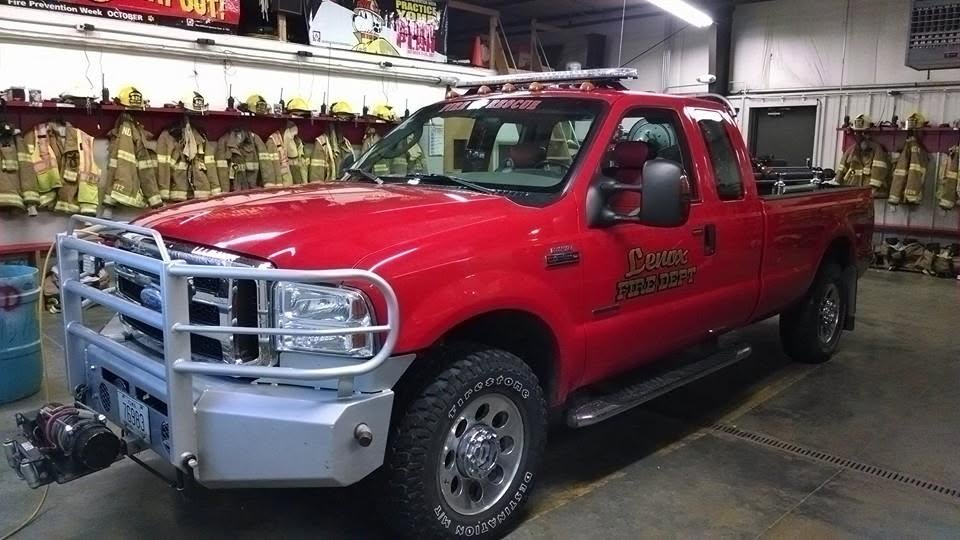 Red Lenox Fire Department truck parked inside fire station with firefighting gear hanging in background.