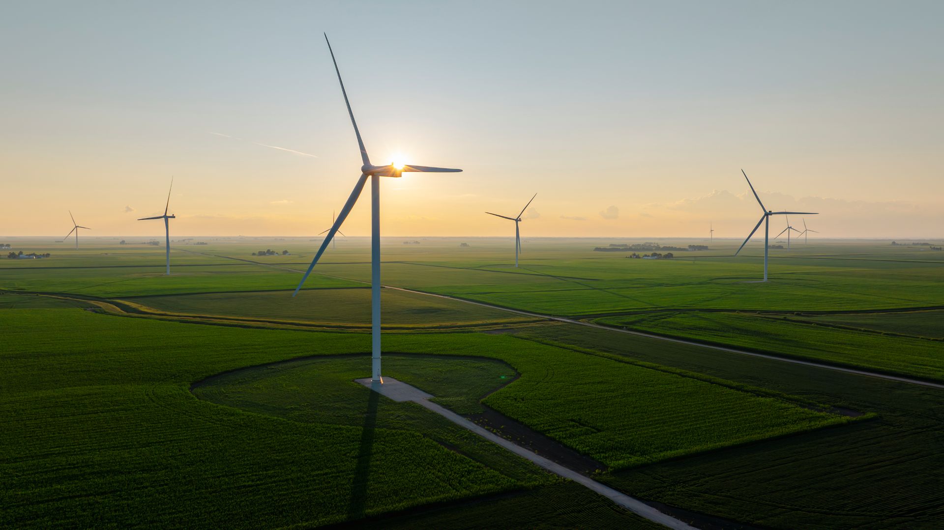 Wind turbines scattered across vast green fields at sunset with the sun behind a turbine blade.