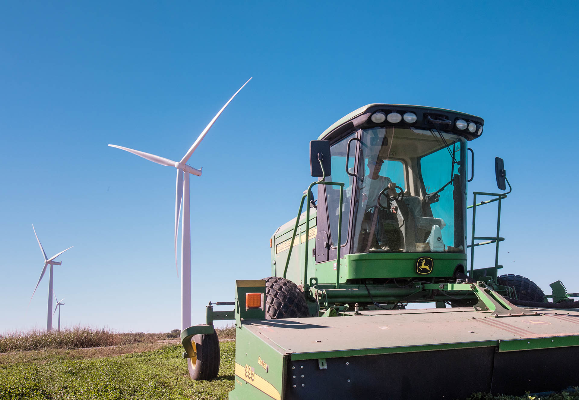 Green John Deere agricultural machine in a field with wind turbines against a clear blue sky.