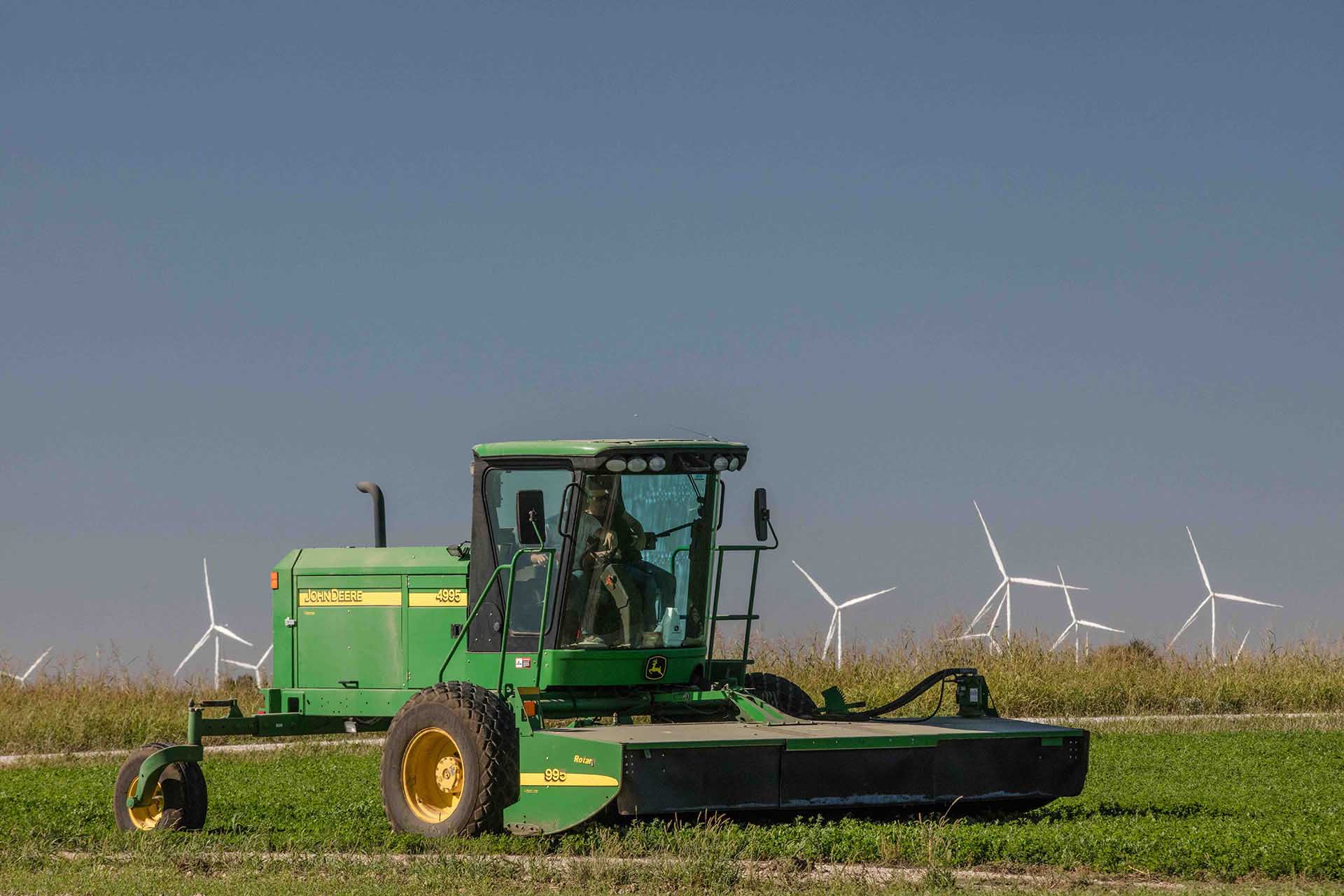 Green John Deere agricultural machine in a field with wind turbines against a clear blue sky.