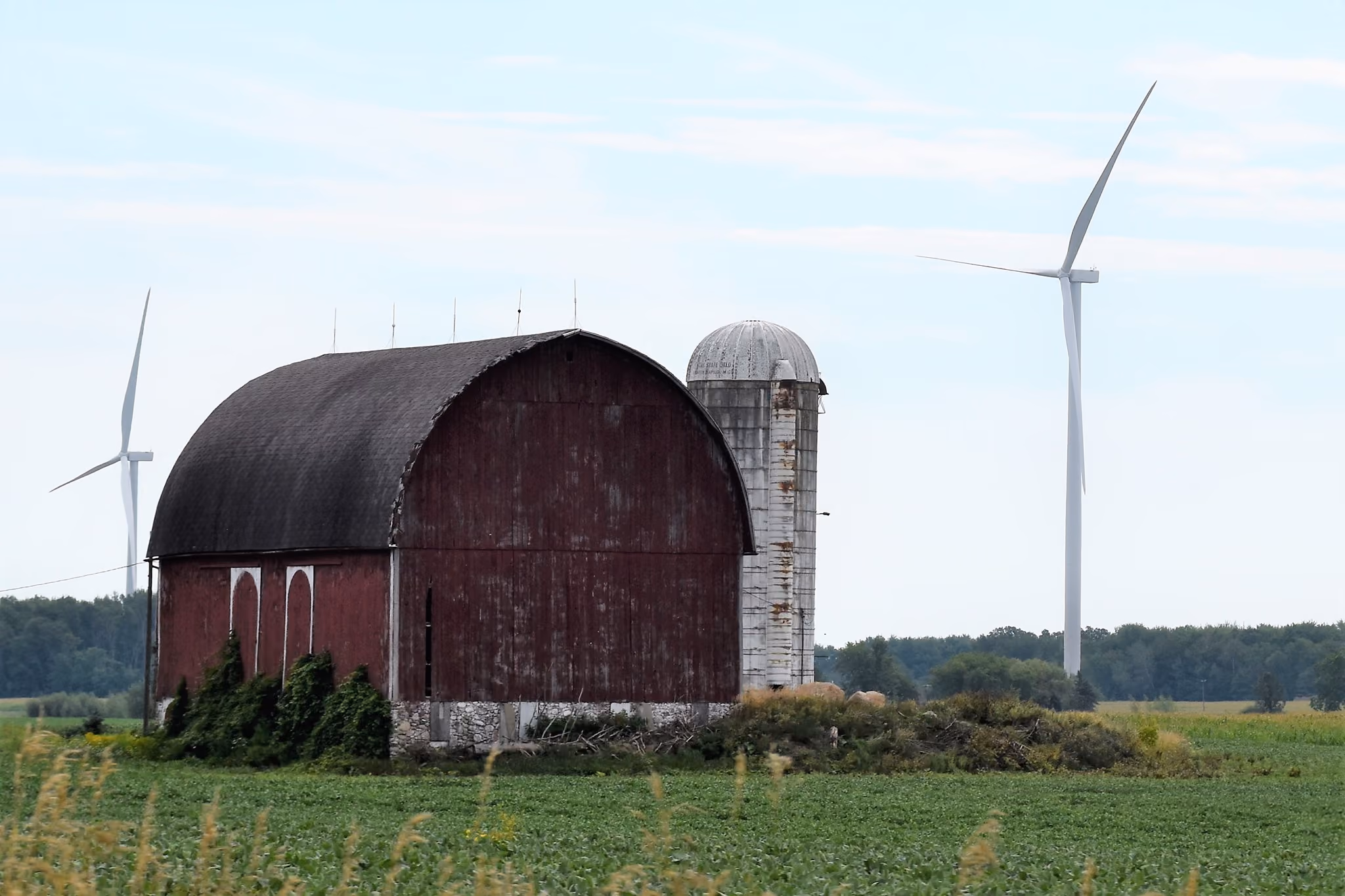 Old red barn and weathered silo in green field with two large white wind turbines behind them.