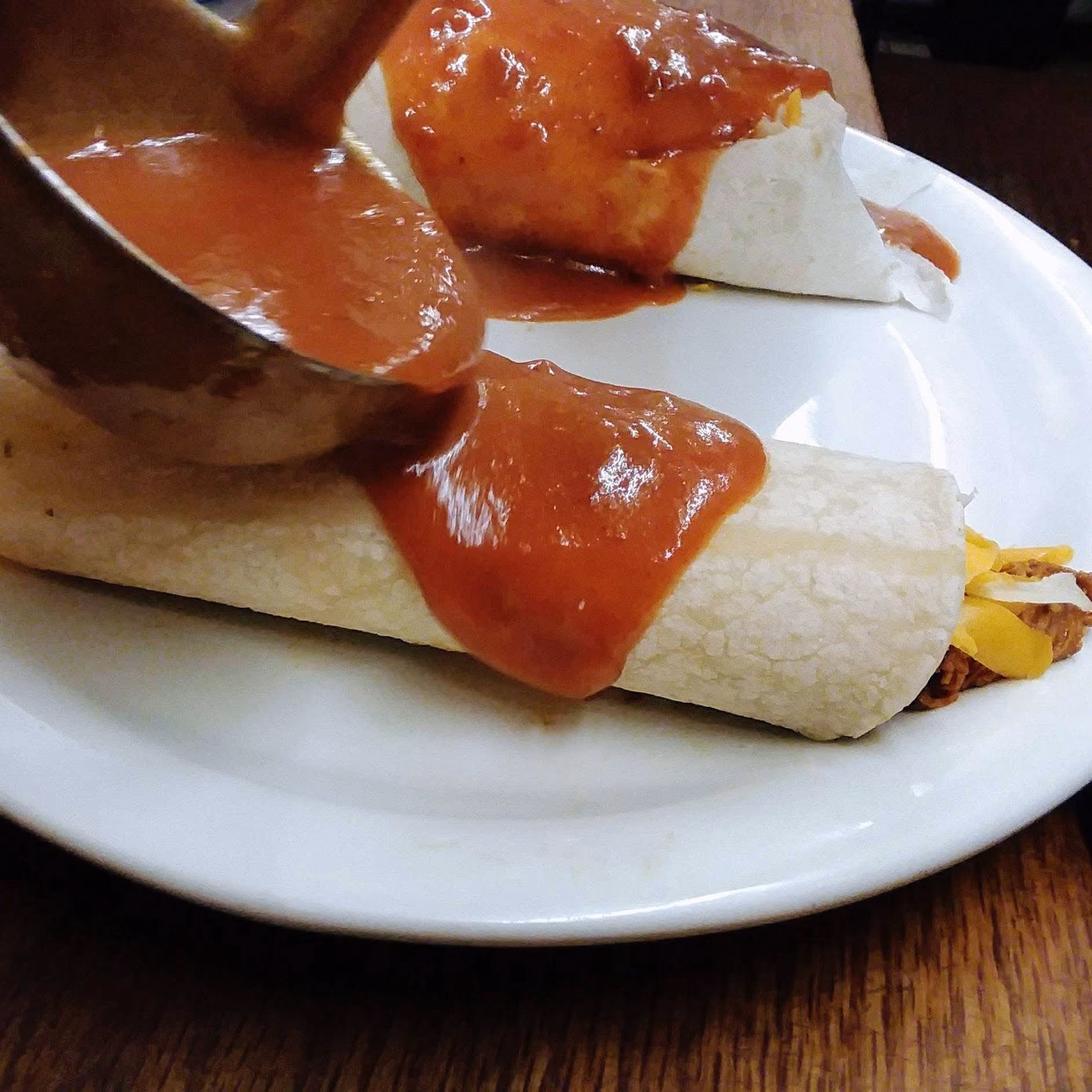 Two rolled tortillas on a white plate; red sauce is being poured over one tortilla from a ladle.