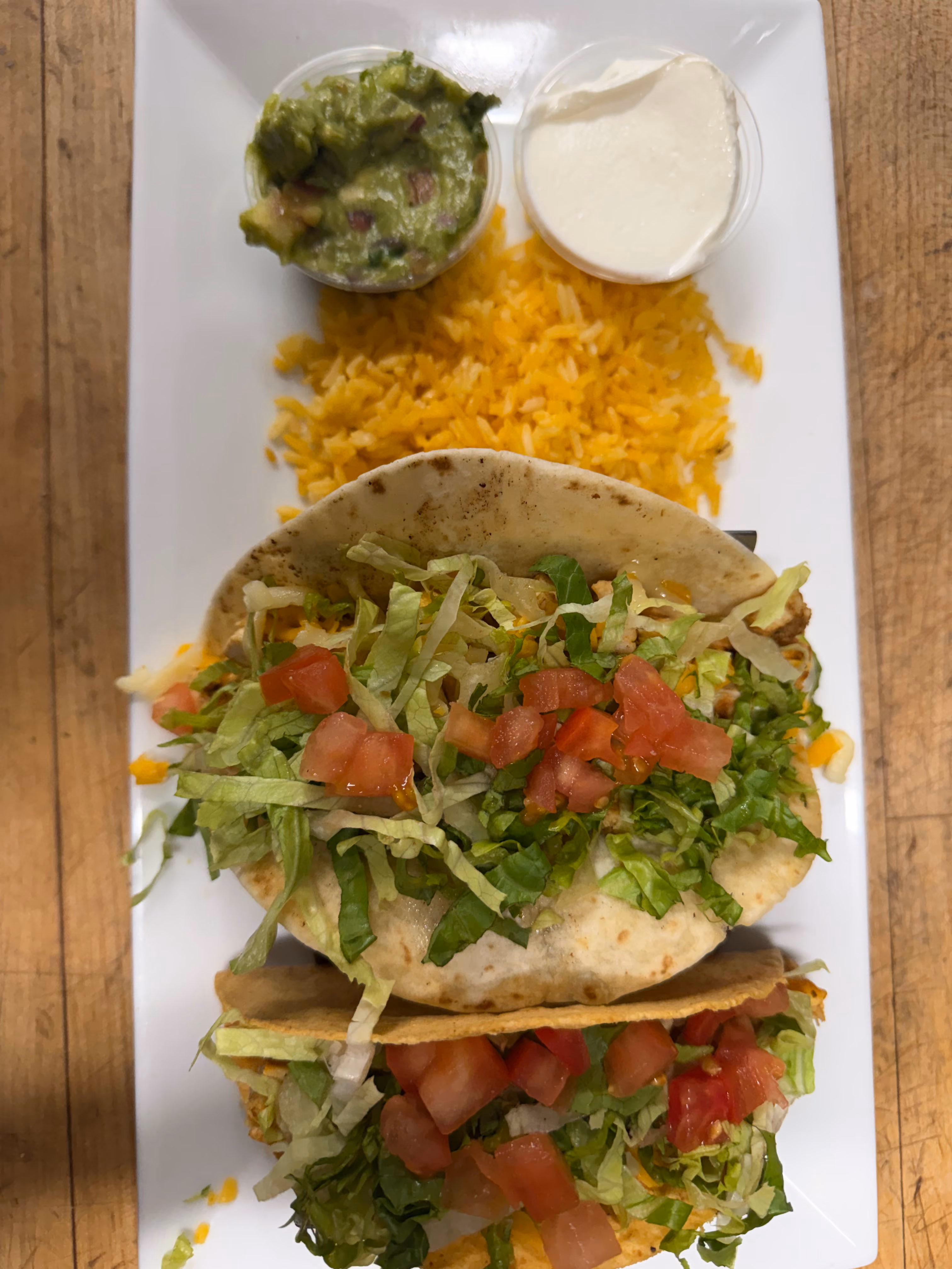 Plate with two tacos filled with lettuce and diced tomatoes, served with shredded yellow cheese, a cup of guacamole, and a cup of sour cream.
