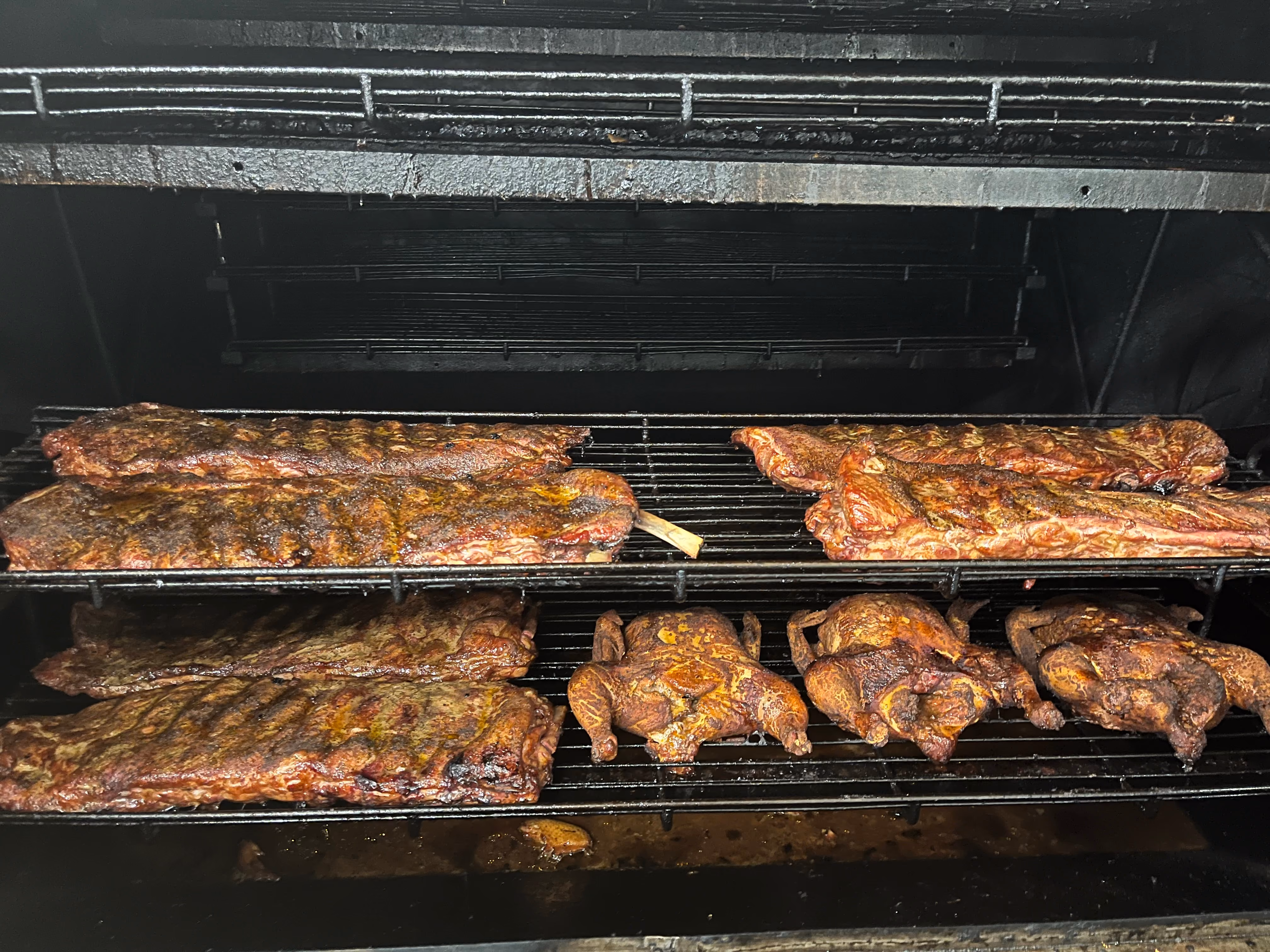 Smoked ribs and whole chickens cooking on metal racks inside a large smoker.