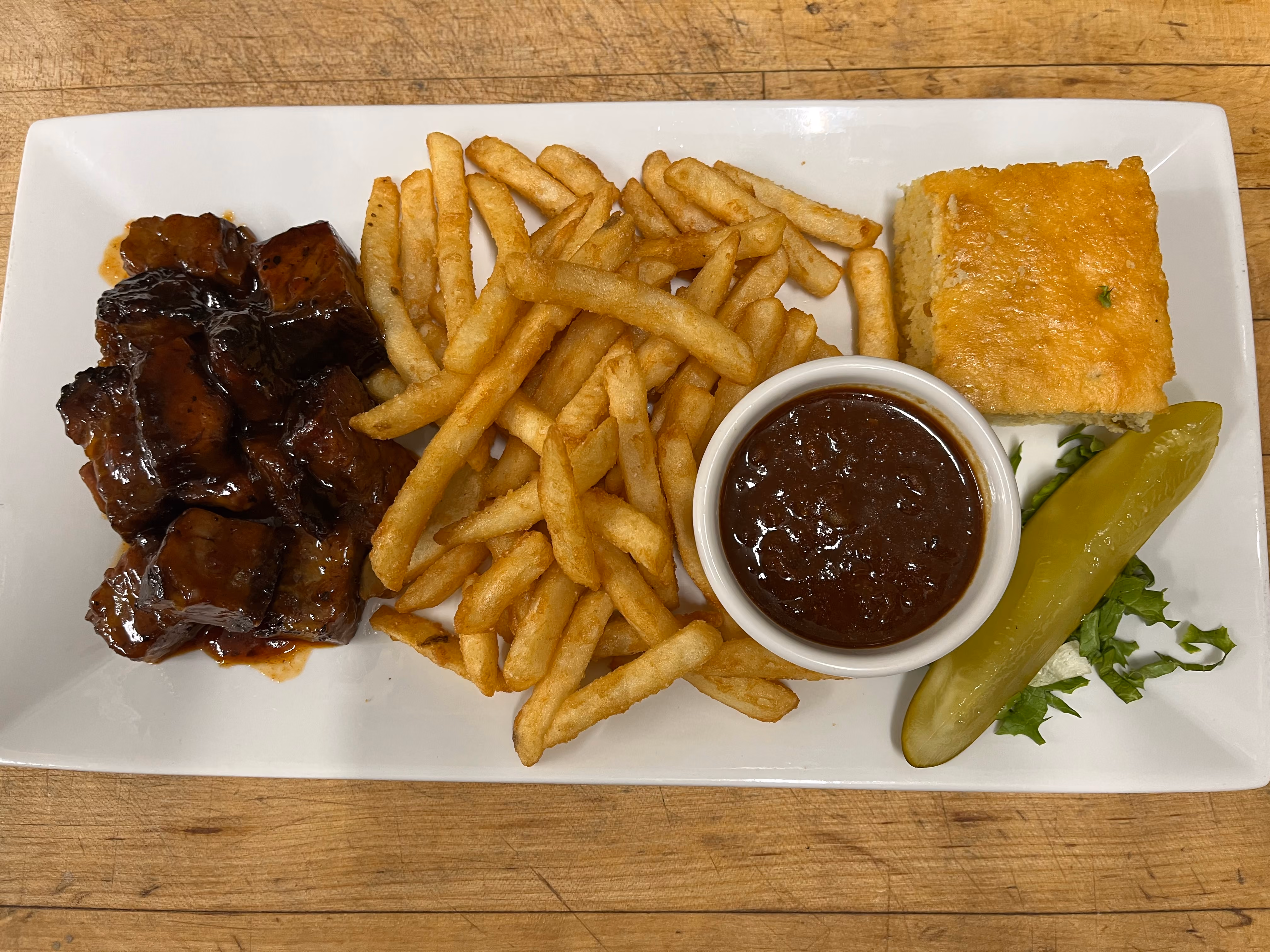 Plate with barbecue glazed pork burnt ends, golden french fries, a cup of barbecue sauce, cornbread square, and a pickle spear on shredded lettuce.