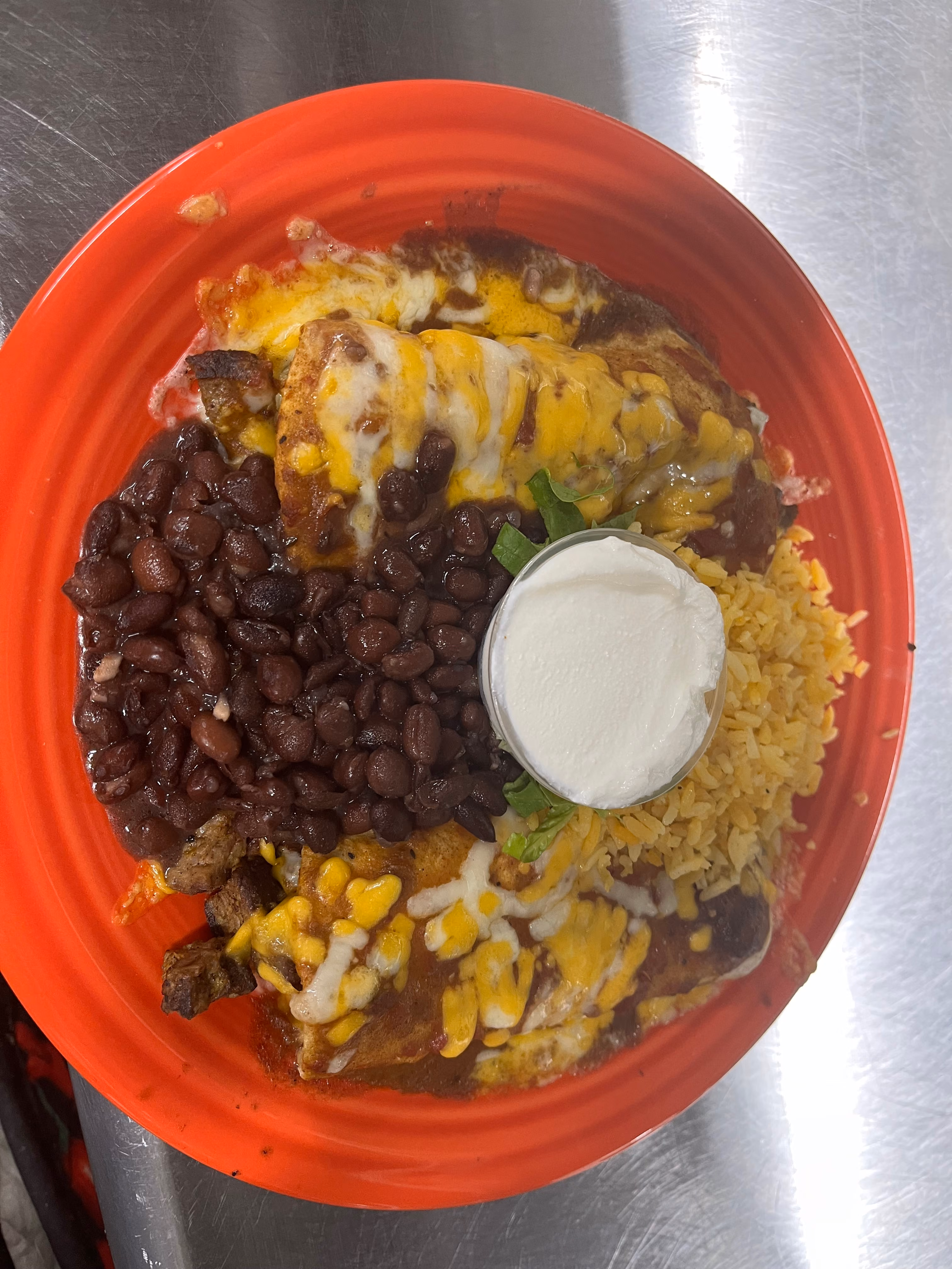 Red plate with enchiladas topped with melted cheese and black beans, rice, and a small cup of sour cream on a metal surface.