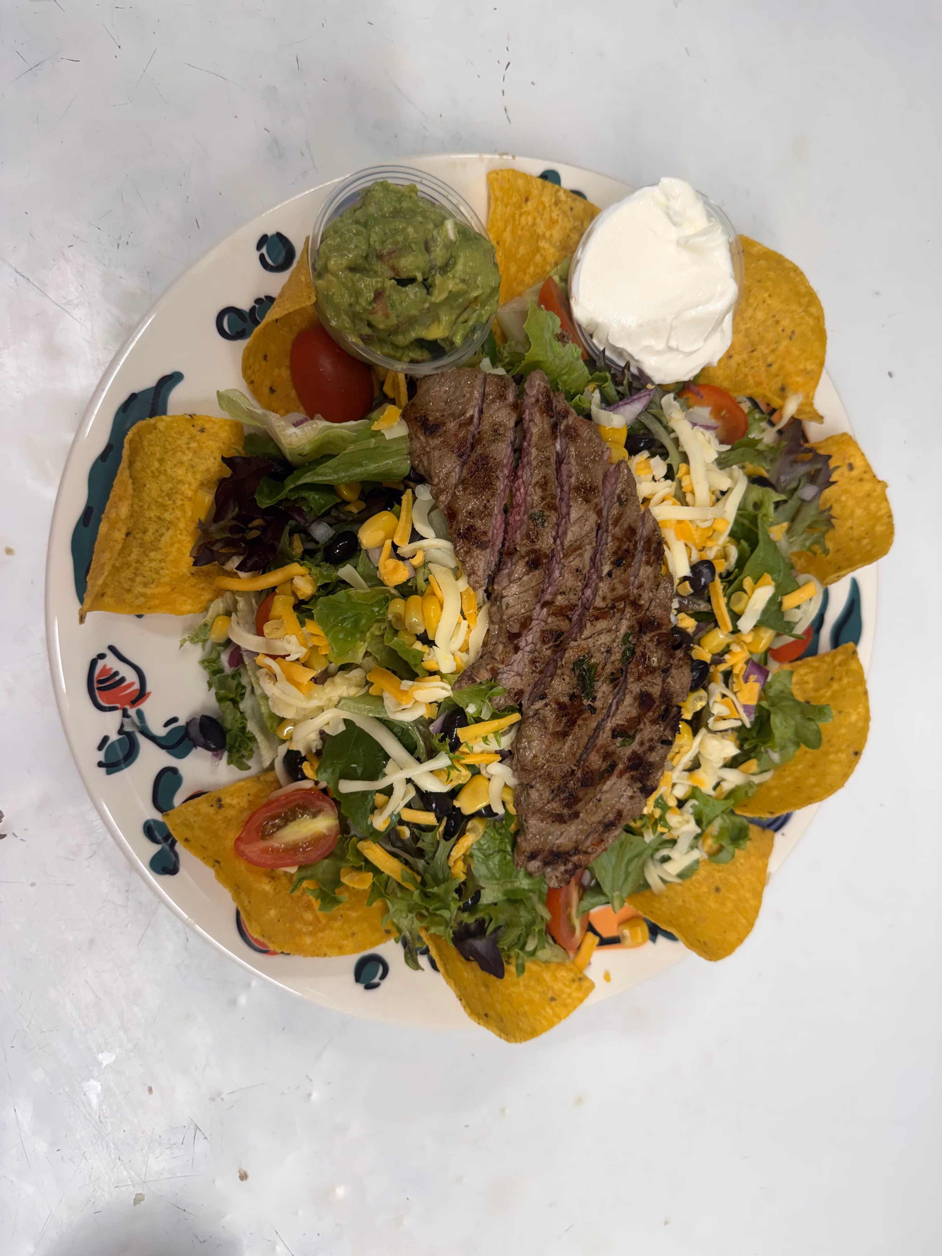 A plate with a grilled steak on top of a fresh salad with mixed greens, shredded cheese, cherry tomatoes, black olives, and surrounded by tortilla chips, served with guacamole and sour cream.