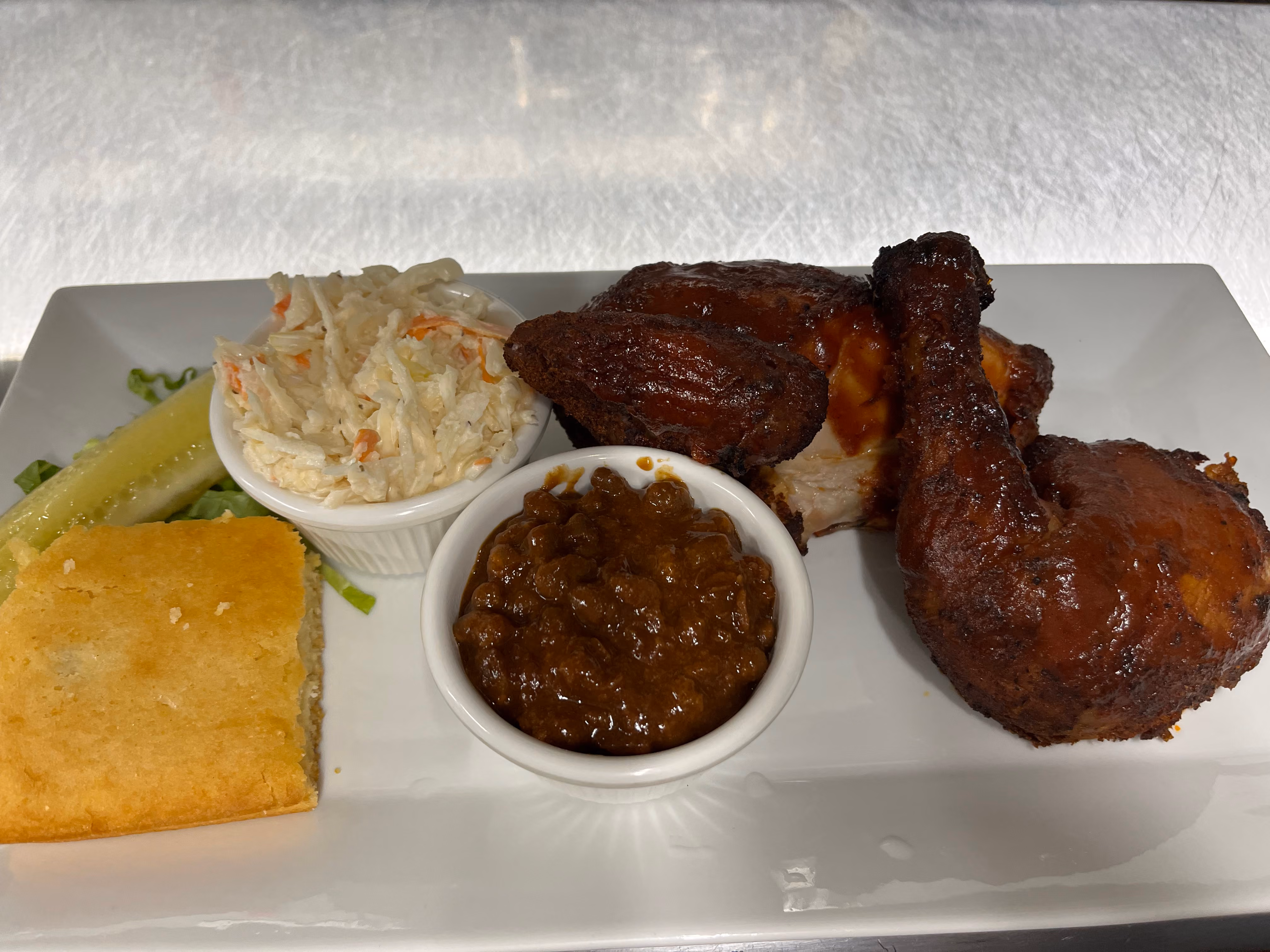 Plate with two pieces of barbecue chicken, a serving of baked beans and coleslaw in small white bowls, a pickle spear, and a piece of cornbread on a white rectangular plate.