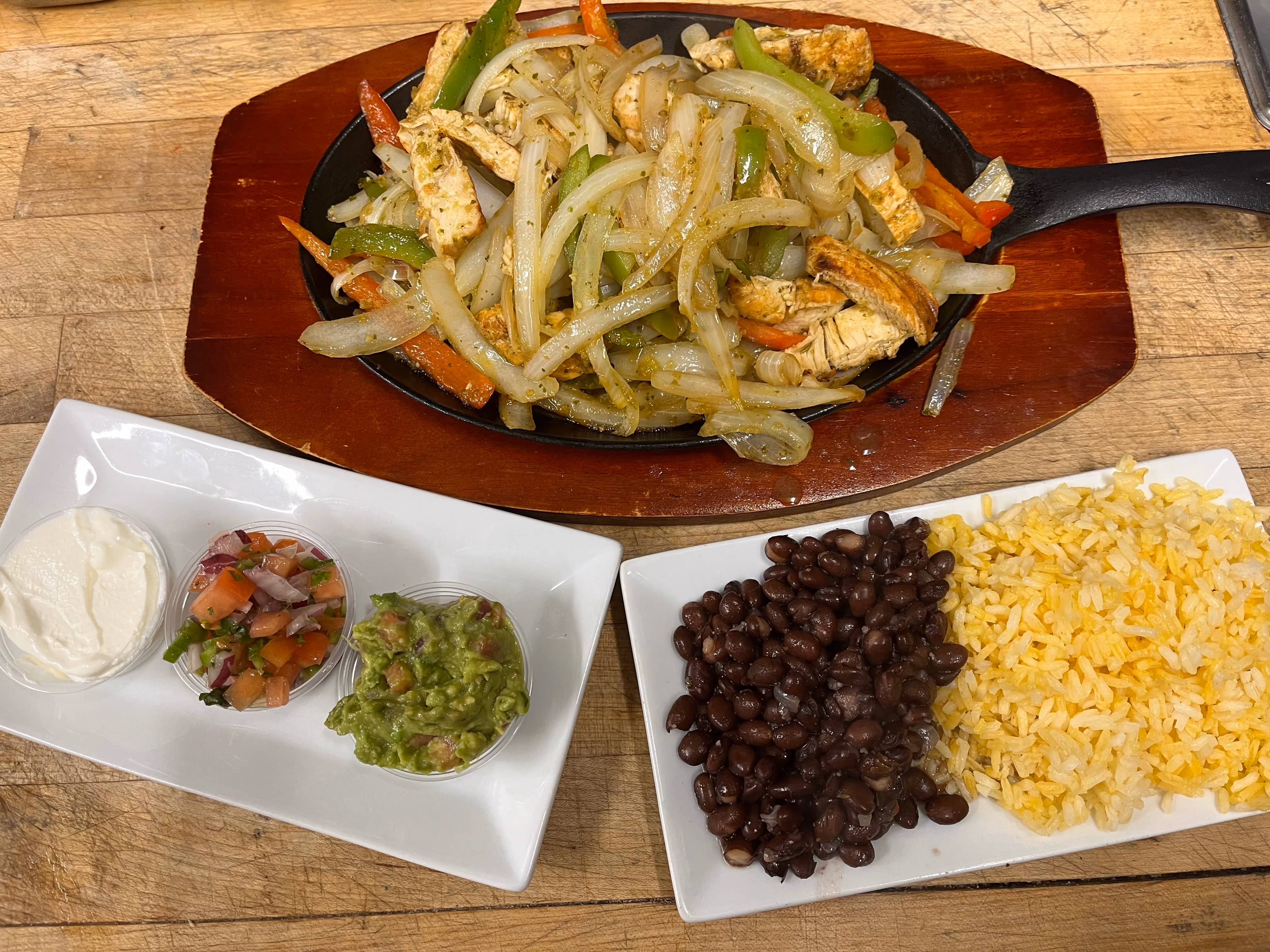 Plate of grilled chicken fajitas with sautéed onions, green and red bell peppers on a wooden platter, served with side dishes of sour cream, pico de gallo, guacamole, black beans, and yellow rice on white rectangular plates.