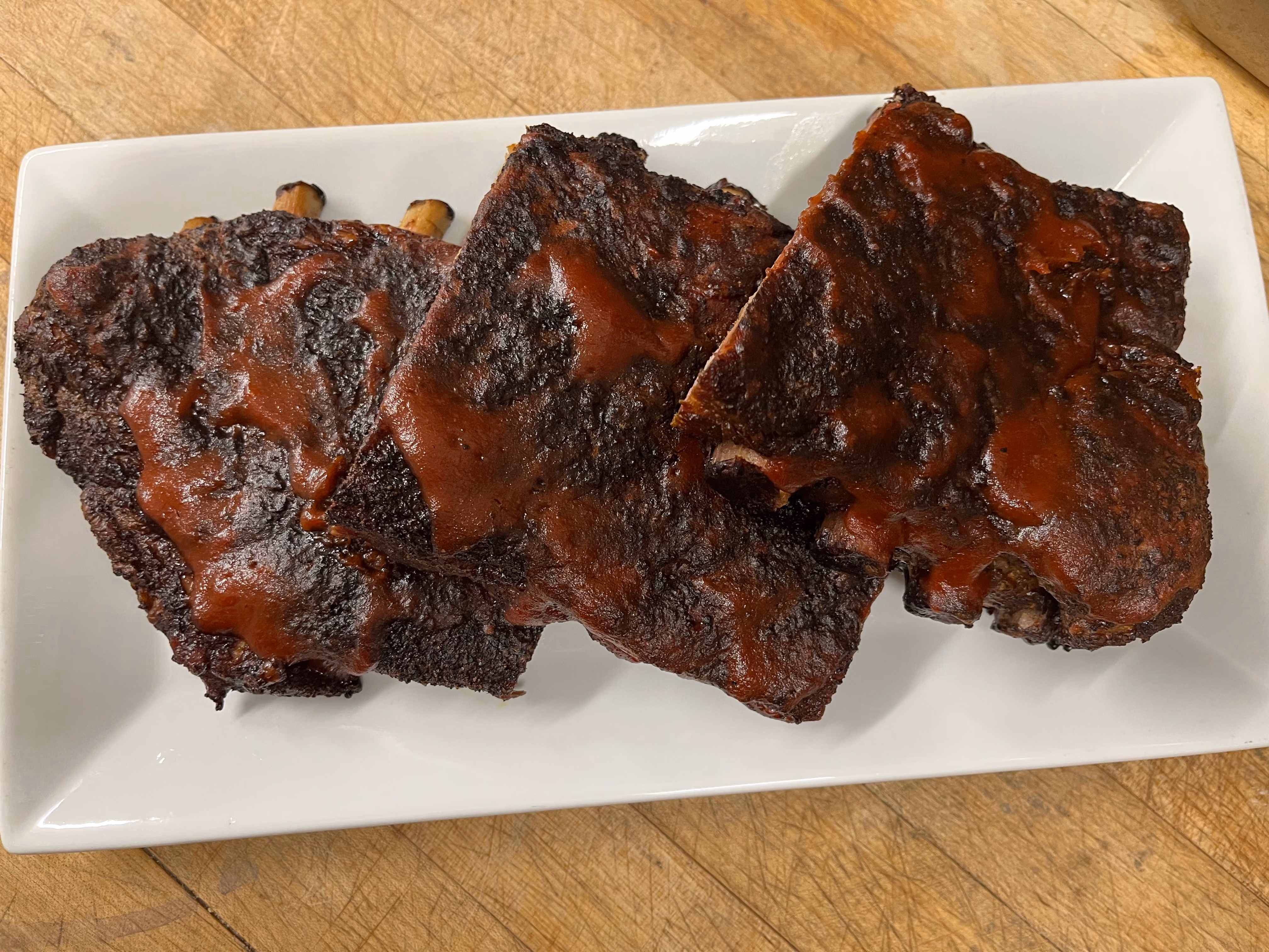 Four pieces of barbecue ribs with smoky char and sauce on a white rectangular plate on a wooden surface.