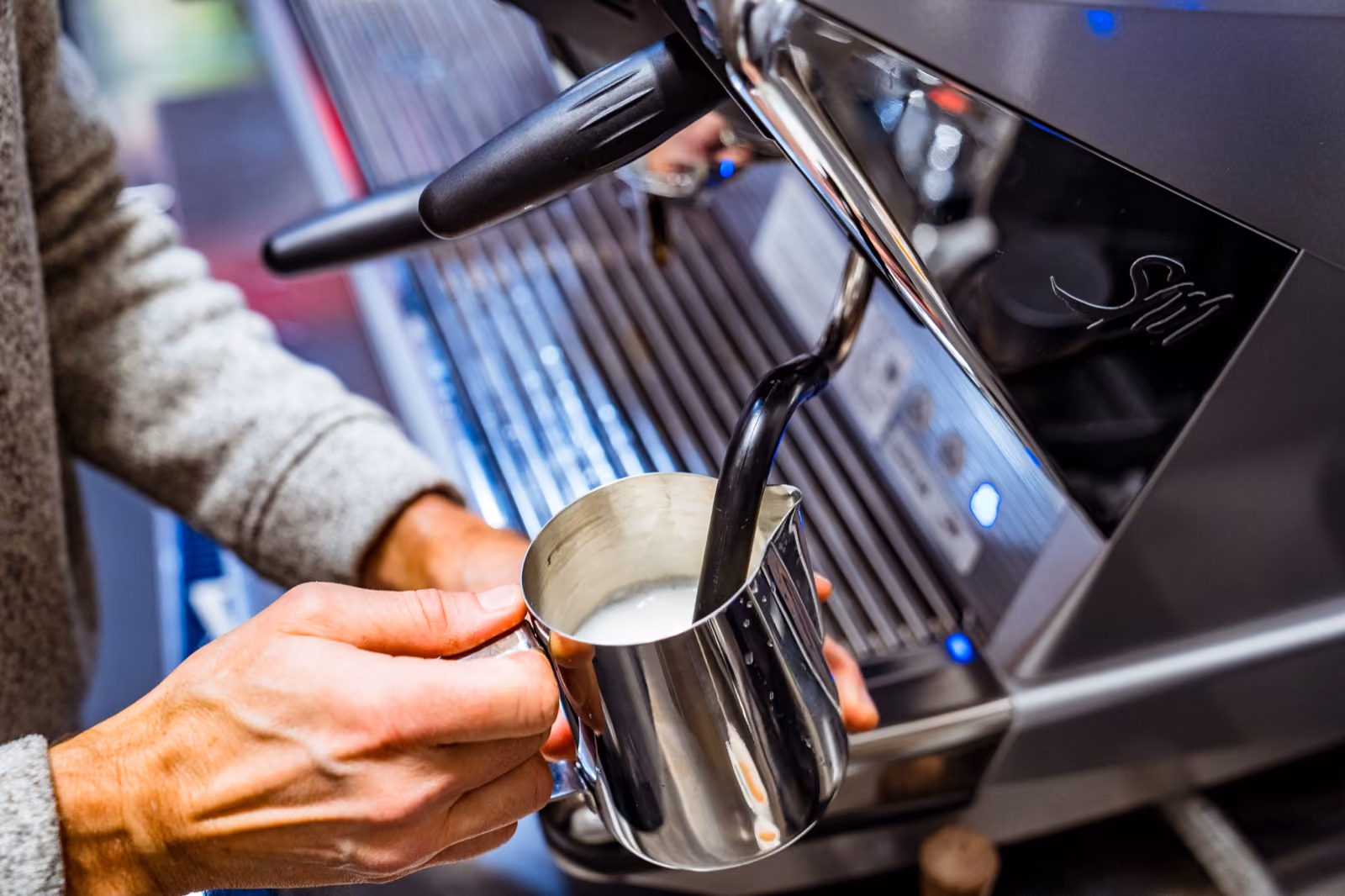 A barista steaming milk in a stainless pitcher at an espresso machine