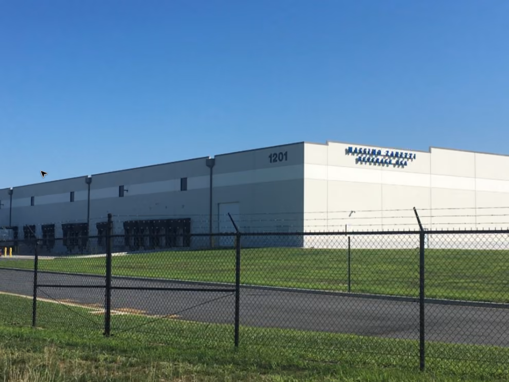 A large industrial warehouse building with the number 1201 on the corner under a clear blue sky, surrounded by a chain-link fence topped with barbed wire and green grass.