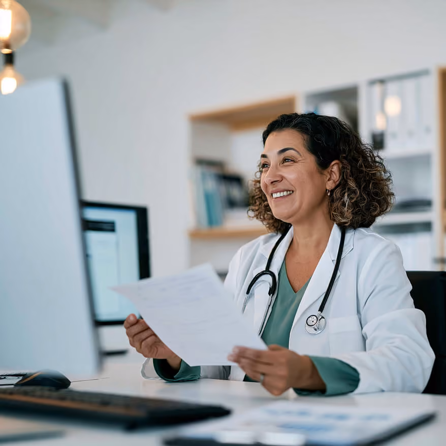 Smiling female doctor in a white coat with a stethoscope around her neck, holding a document and sitting at a desk with a computer in a medical office.