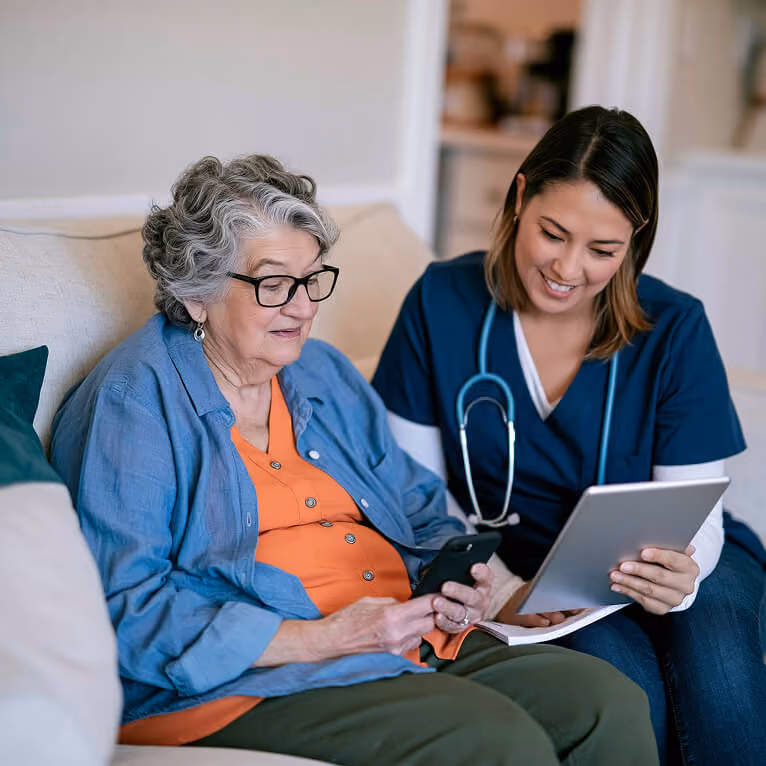 Senior woman sitting on a couch with a younger female healthcare professional who is showing her something on a tablet while the woman holds a smartphone.
