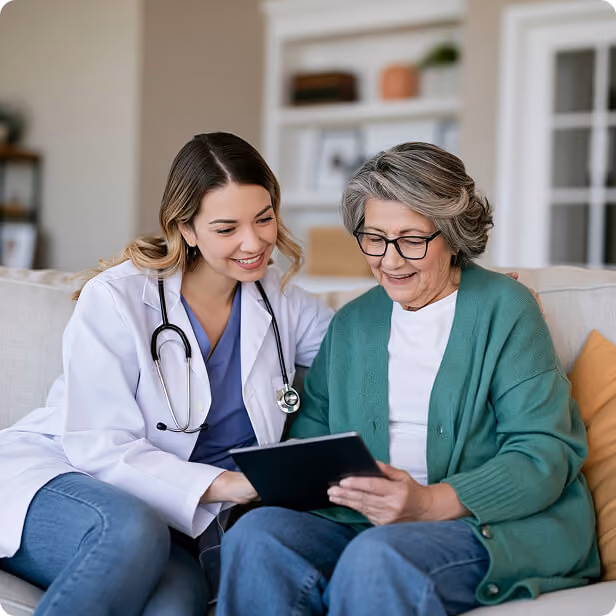 A female doctor with a stethoscope around her neck sitting on a couch next to an elderly woman with gray hair and glasses who is holding and looking at a tablet, both smiling.