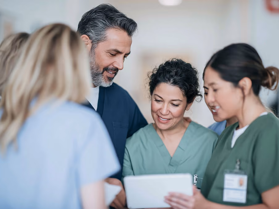 A group of diverse healthcare professionals in scrubs gathered together, smiling and reviewing information on a digital tablet.