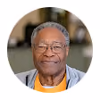 Smiling elderly Black man wearing glasses, a gray jacket, and an orange shirt against a blurred indoor background.