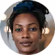 Portrait of a woman with dark skin and braided hair styled with a dark blue headband, looking directly at the camera with a neutral expression, with blurred background.