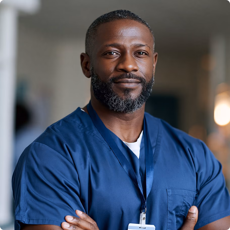 Confident man in blue medical scrubs with ID badge standing with arms crossed, looking at camera.