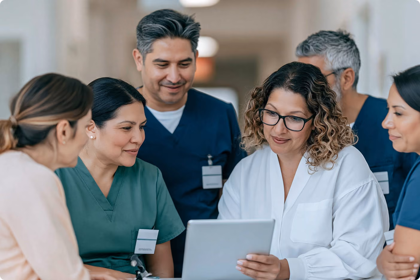 A group of five healthcare professionals, including doctors and nurses in scrubs and lab coats, gathered around a tablet, looking at it attentively in a hospital corridor.