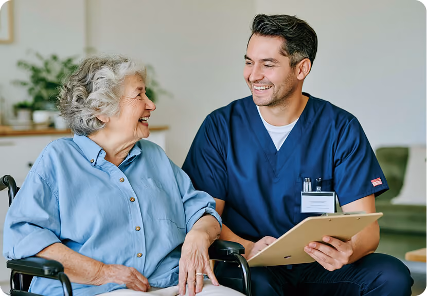 Smiling male healthcare worker in navy scrubs holding a clipboard while talking with a happy elderly woman seated in a wheelchair.