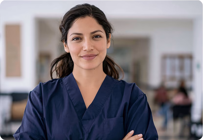 Confident woman in navy blue medical scrubs with arms crossed, smiling slightly in a bright, blurred indoor setting.
