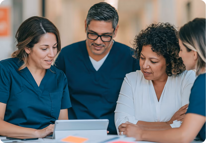 Four healthcare professionals, three in blue scrubs and one in white, gathered around a tablet device discussing or reviewing information.