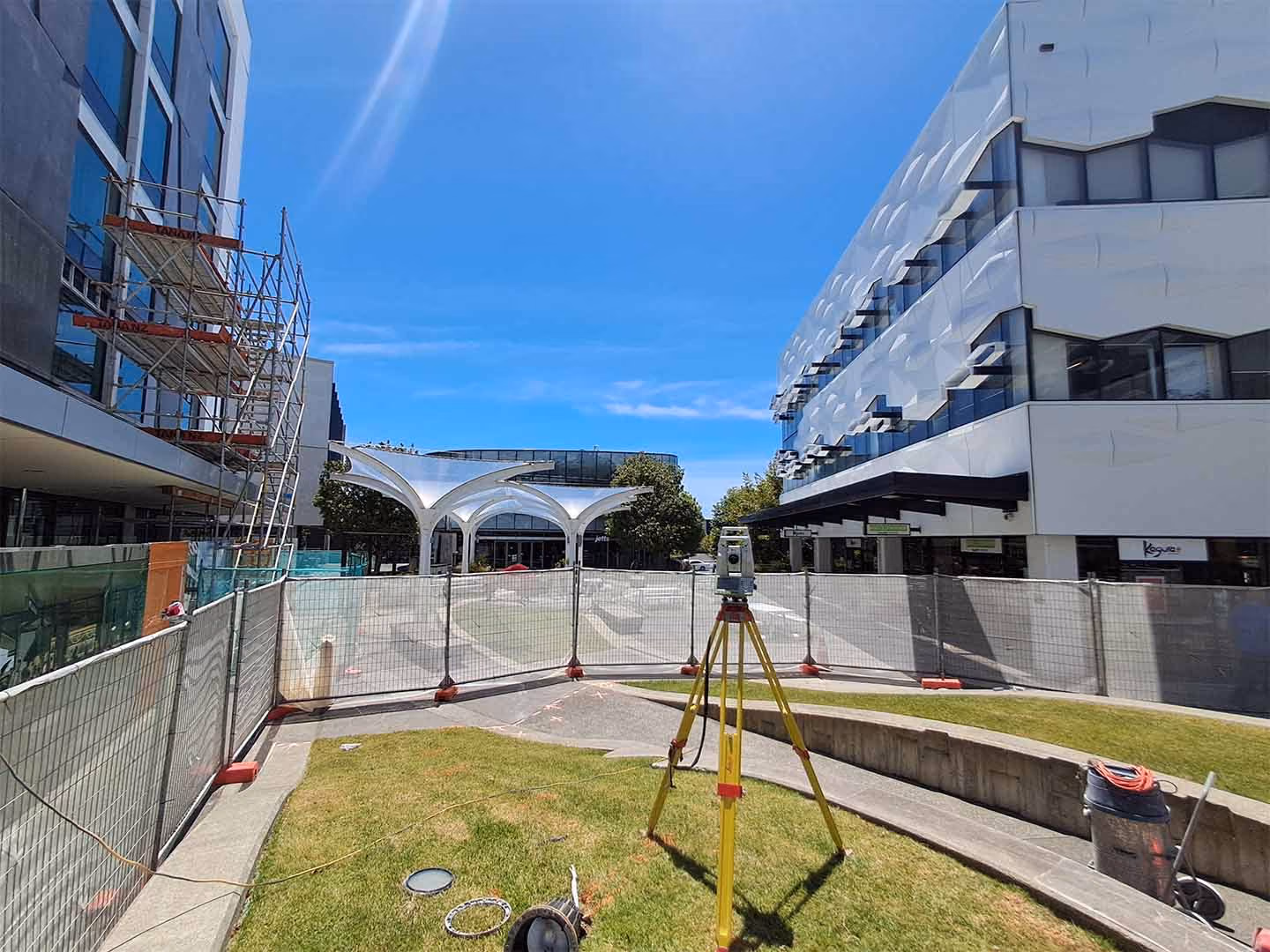 Construction site with metal fencing, a surveyor tripod, and modern buildings under a clear blue sky.