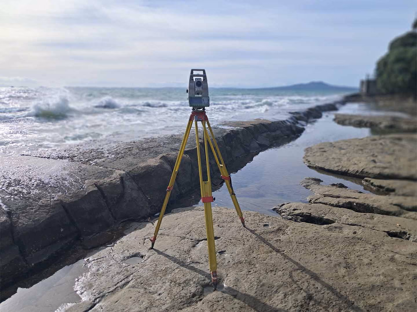 Surveying tripod with equipment set up on rocky shore near ocean waves and water channel.