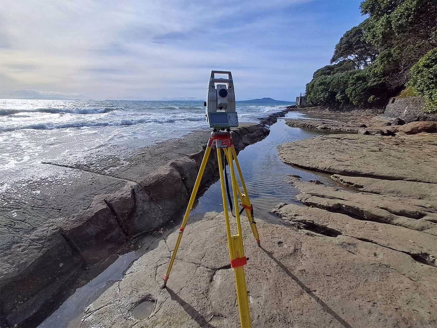 Surveying instrument on a tripod set up on rocky shore with ocean waves and trees in background.
