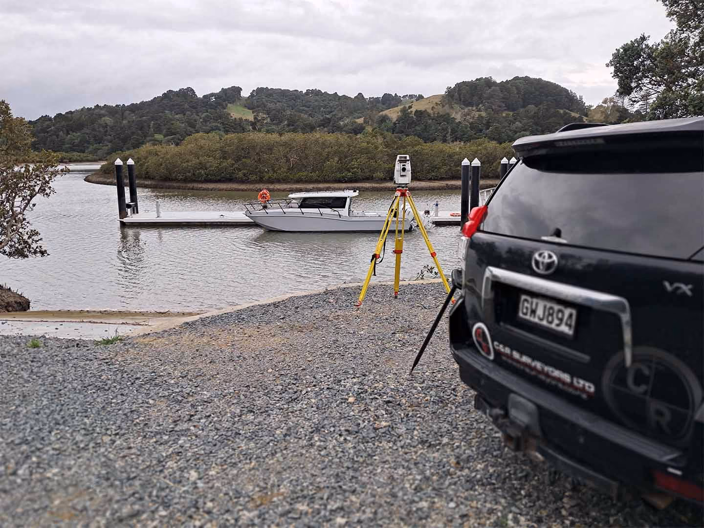 Surveying tripod with equipment on a gravel shore near a boat docked on a river with forested hills.