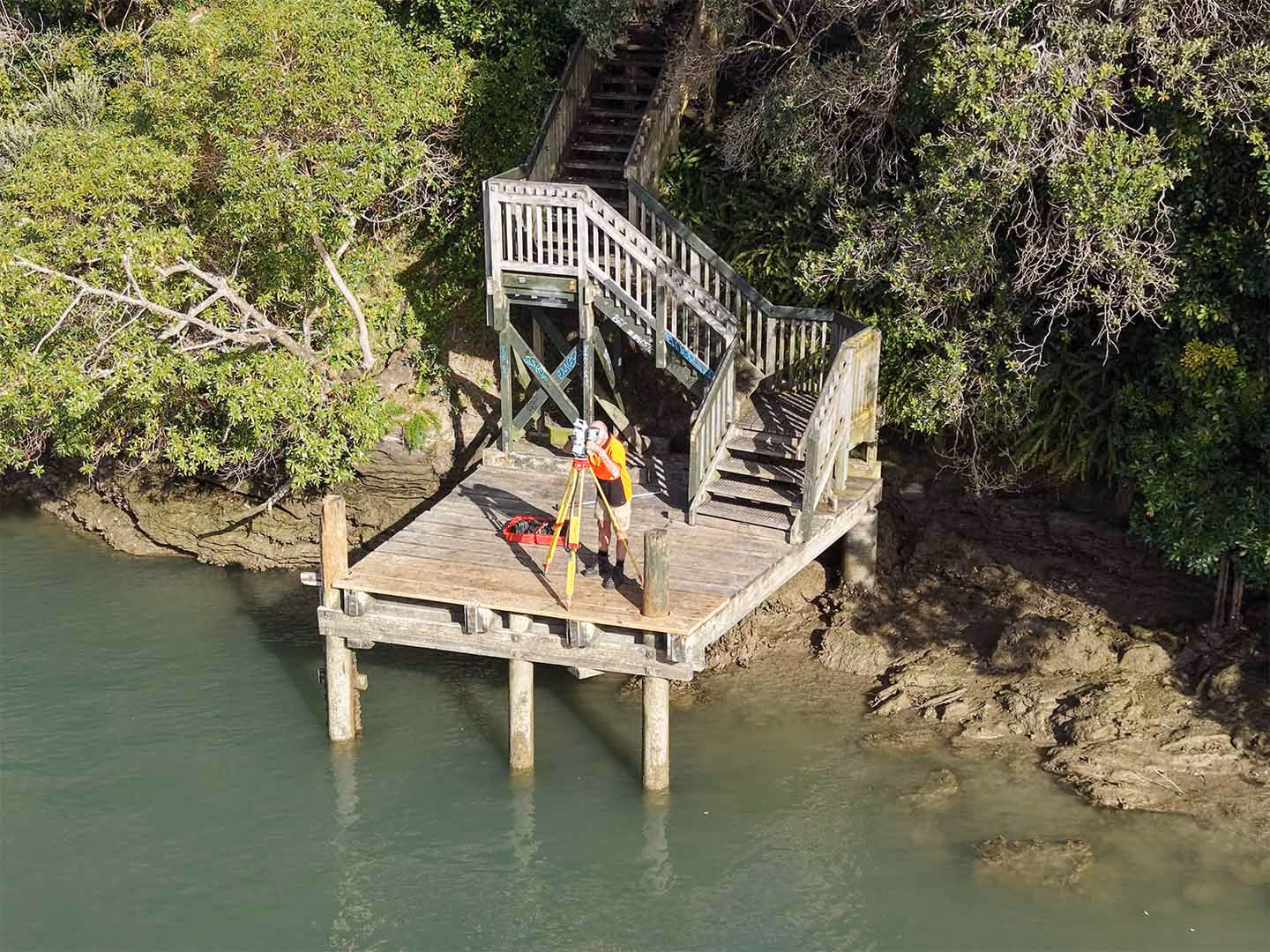 Man with orange shirt using survey equipment on wooden platform by water and stairs in green foliage.