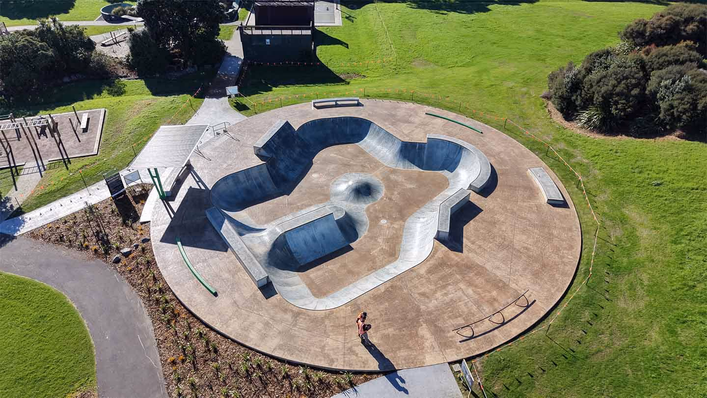 A concrete skatepark with bowls and ramps surrounded by green grass and a person standing nearby.