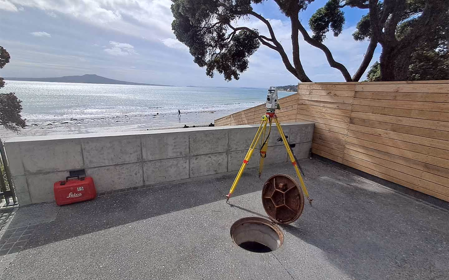 Surveying tripod with equipment near an open manhole by a beach with ocean and distant land.