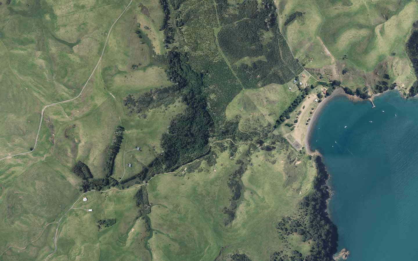 Aerial view of green fields, forest patches, small buildings, and a bay with blue water and boats.