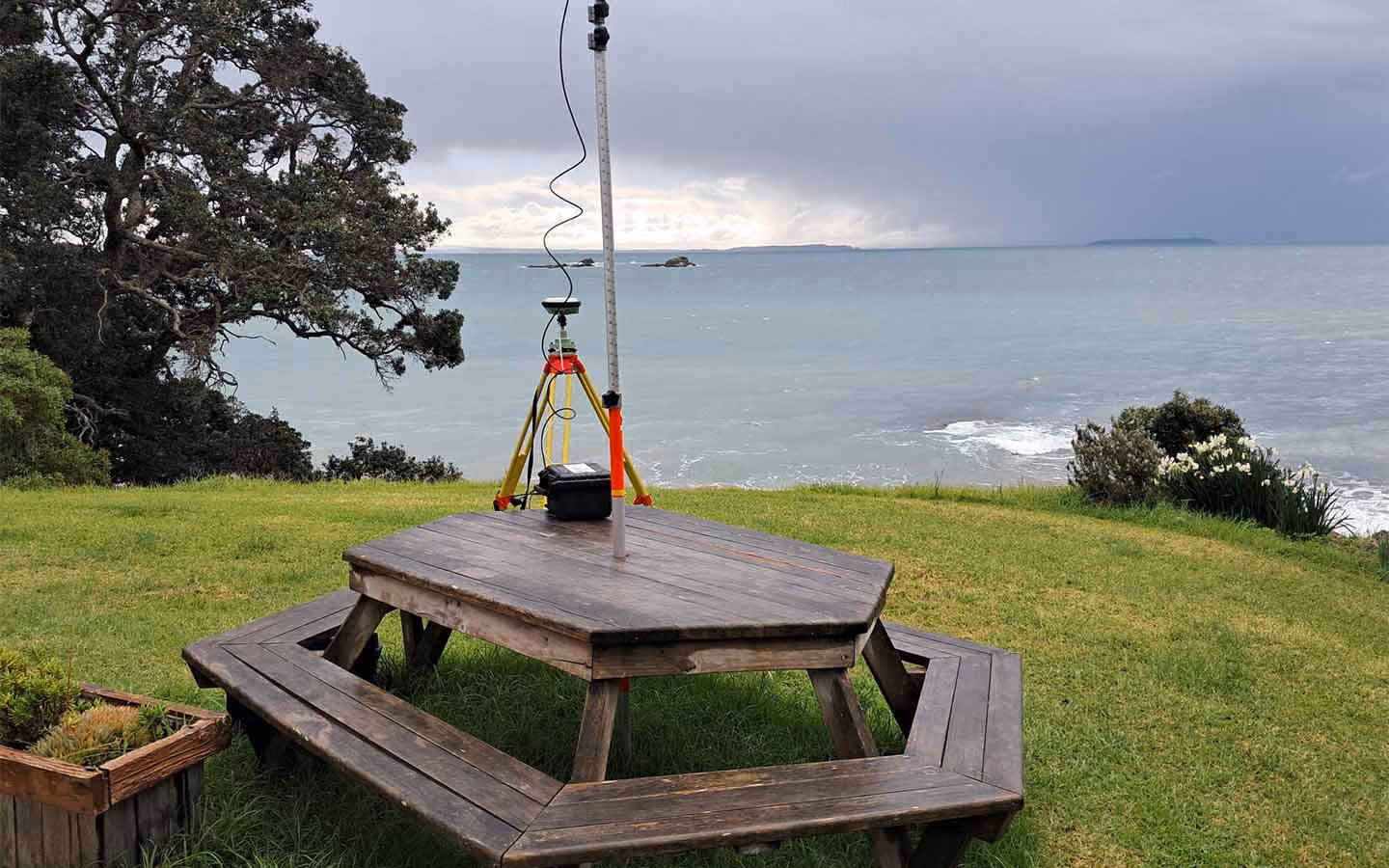 Wooden octagonal picnic table on grass by ocean under cloudy sky with surveying equipment on it.