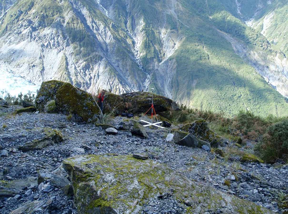 Rocky mountain slope with moss-covered boulders and a surveying tripod near a green valley.