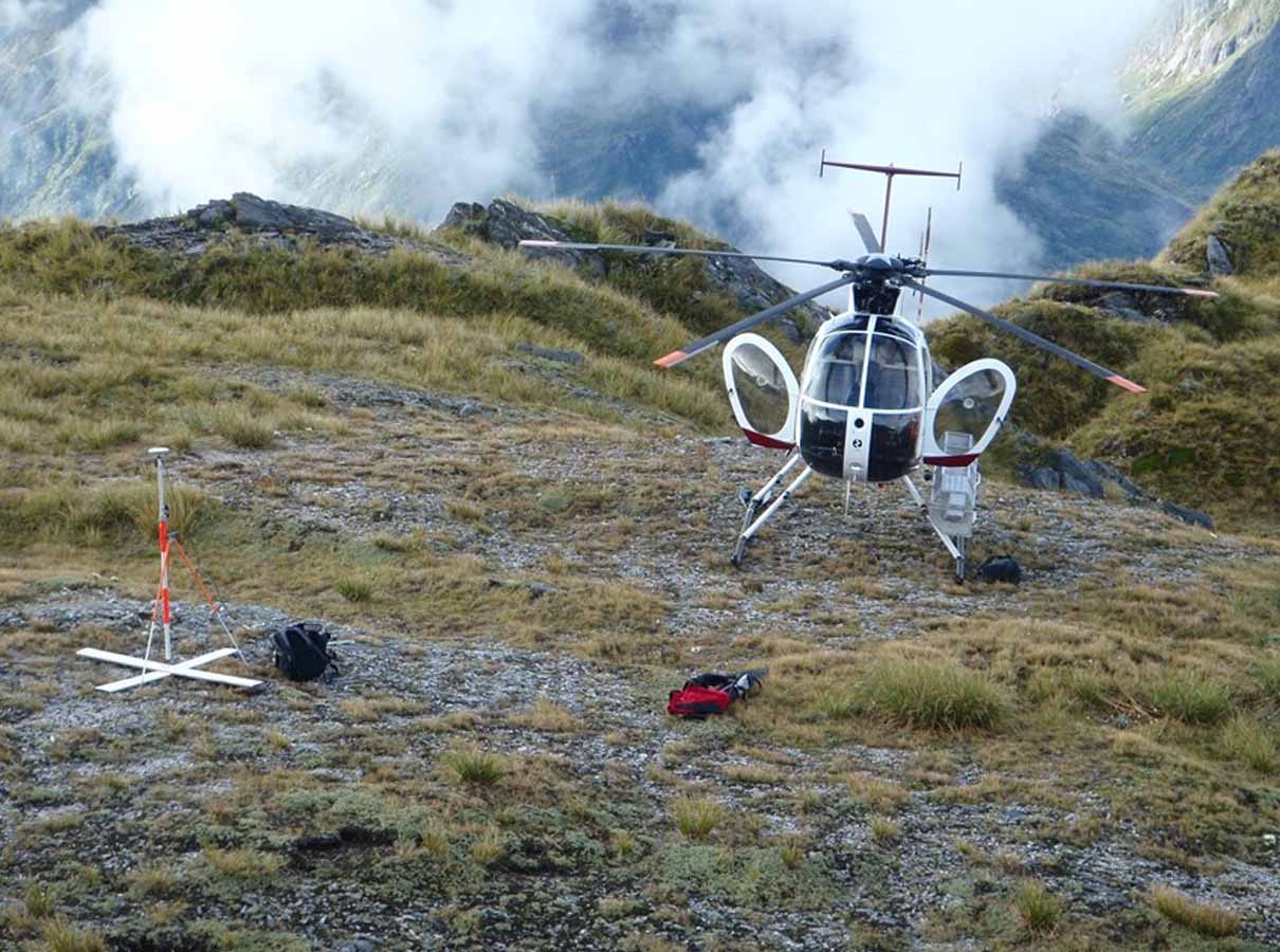 White helicopter with open doors landed on a grassy mountain terrain with equipment nearby.