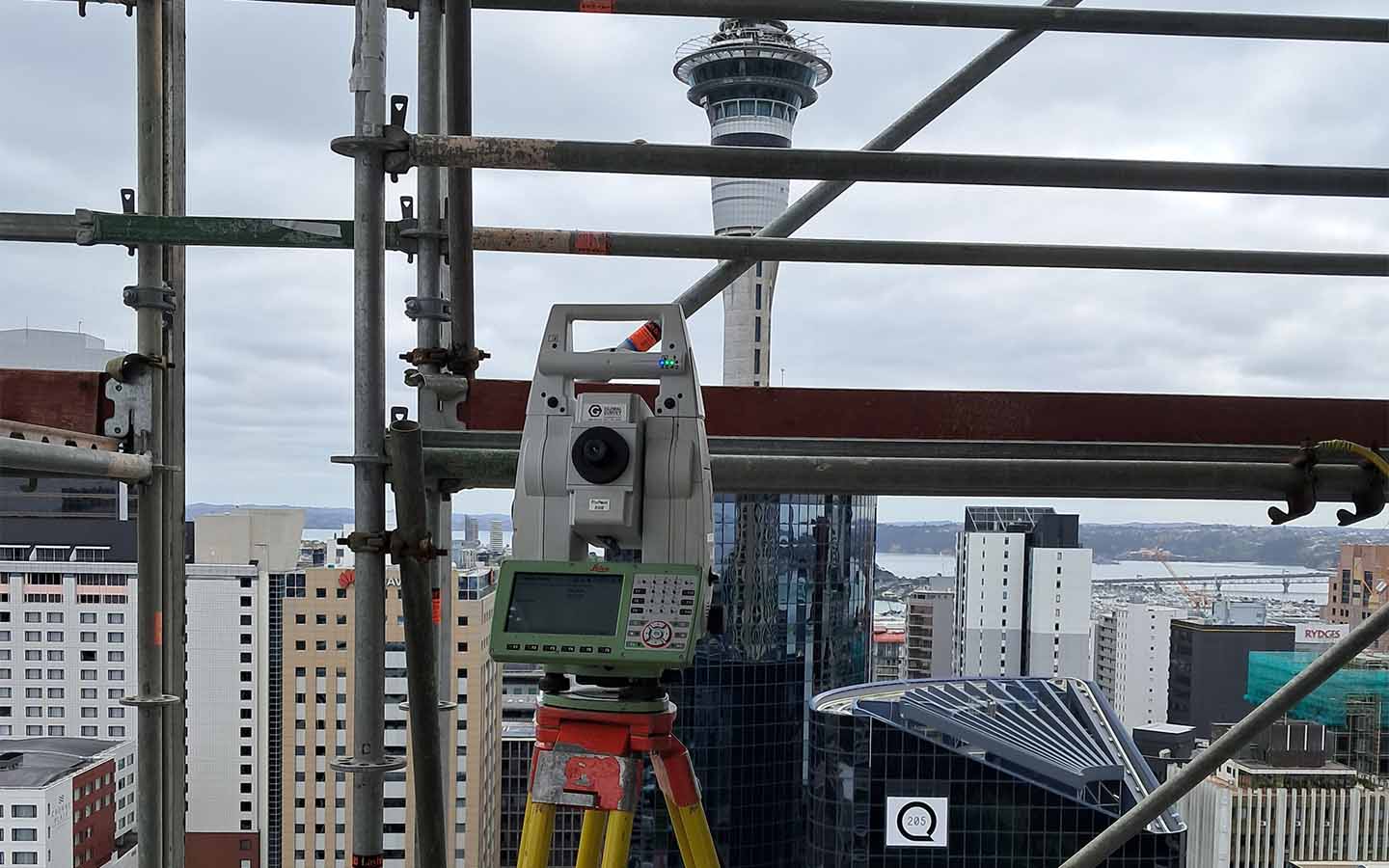 Surveying instrument on tripod within scaffolding against city skyline and tower under cloudy sky.
