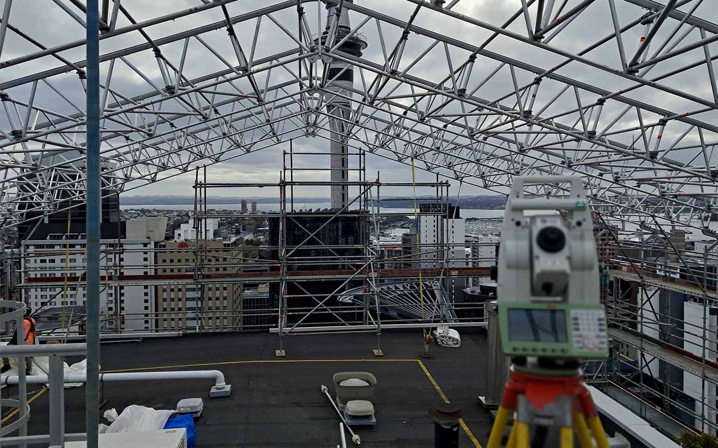 Construction site with metal scaffolding and surveying equipment overlooking city buildings and water in distance.
