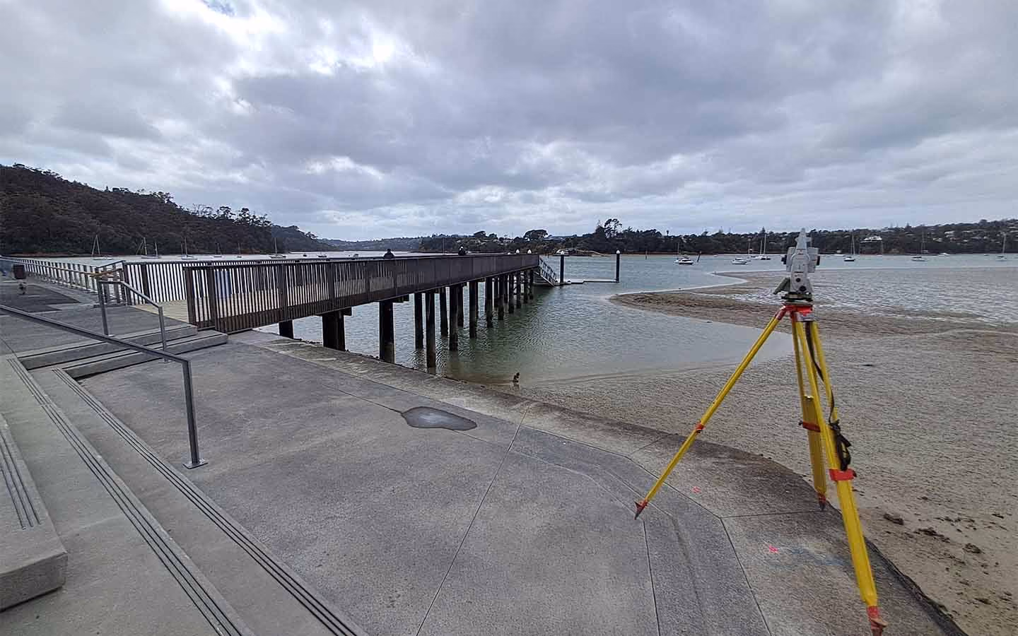 Surveying equipment on tripod by pier extending over water under cloudy sky.