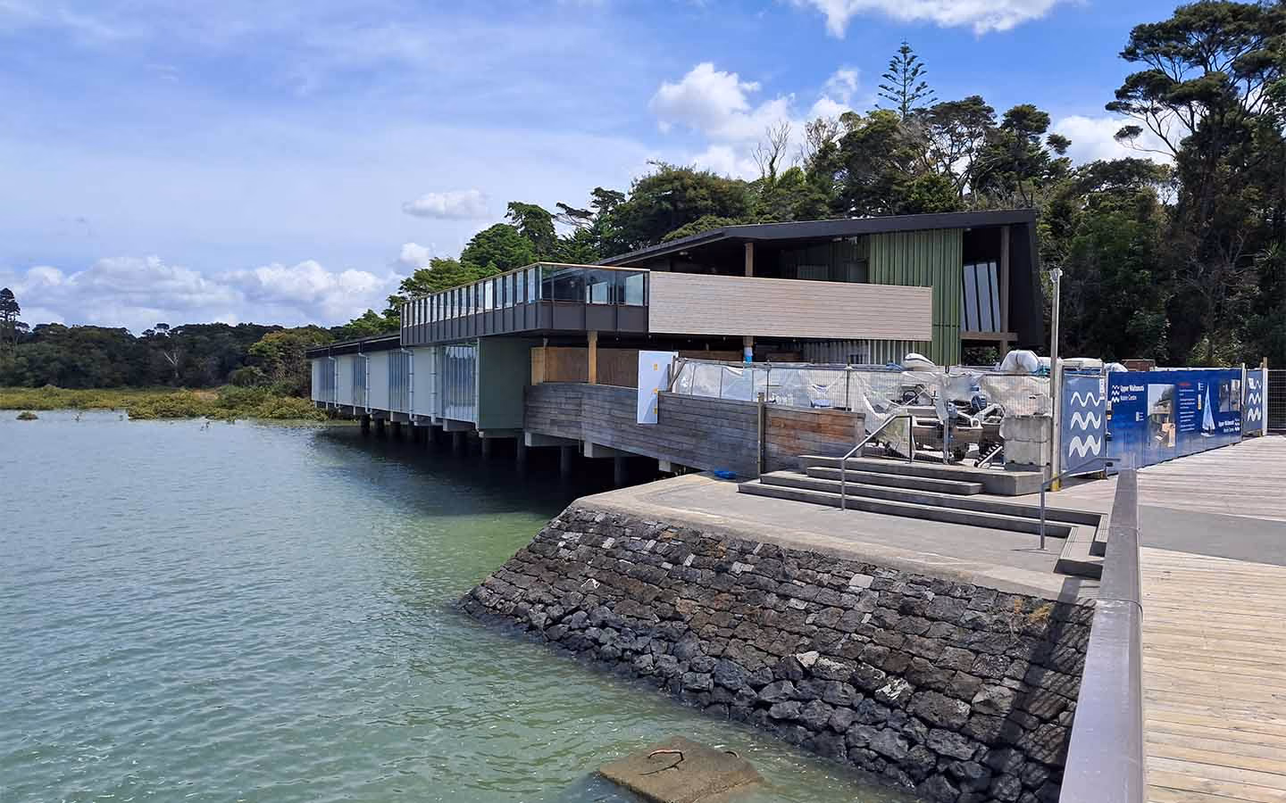 Modern waterfront building on stilts beside a stone embankment and calm water under a partly cloudy sky.