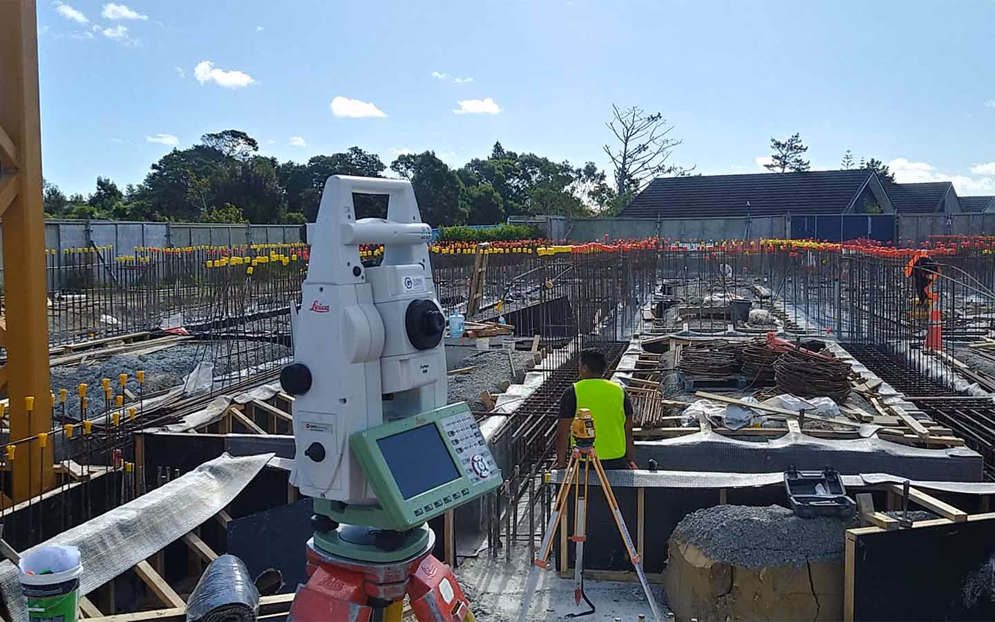 Construction site with a white Leica surveying instrument in the foreground and a worker in a yellow vest.