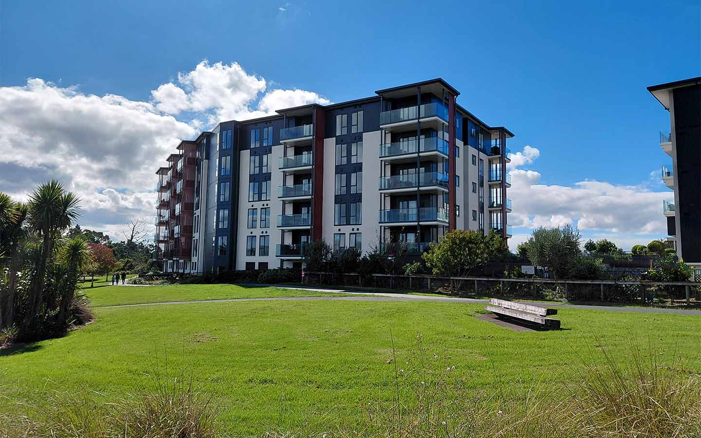 Modern apartment building beside a large green lawn with a bench under a blue sky.