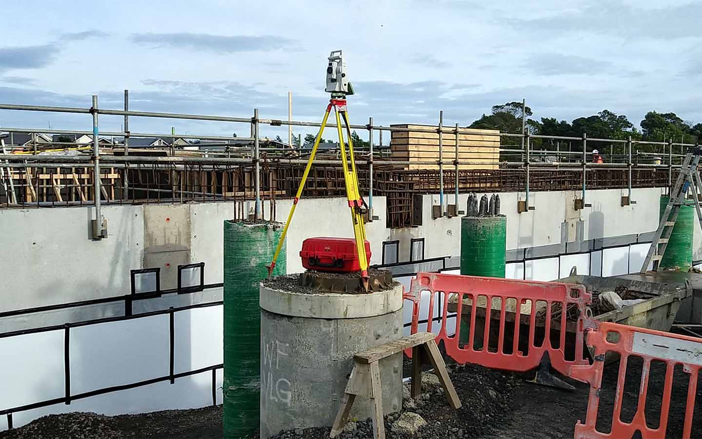 Construction site with concrete pillars, scaffolding, a total station on tripod, and safety barriers.