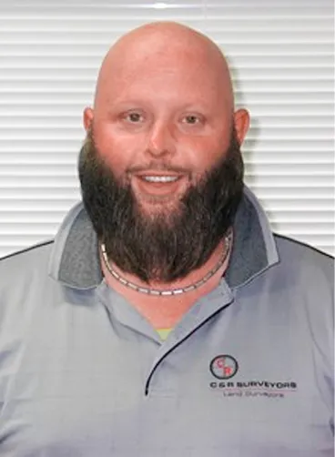 Smiling bald man with a full beard wearing a gray C & R Surveyors polo shirt in front of blinds.