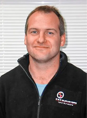 Man with short hair wearing a black zip-up jacket with a surveyors logo stands in front of white blinds.