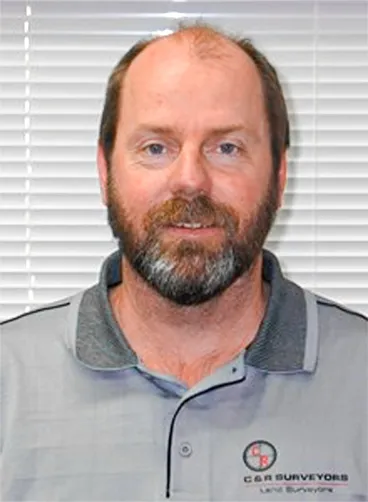 Man with a beard wearing a grey polo shirt with a C.B.S Surveyors logo in front of window blinds.