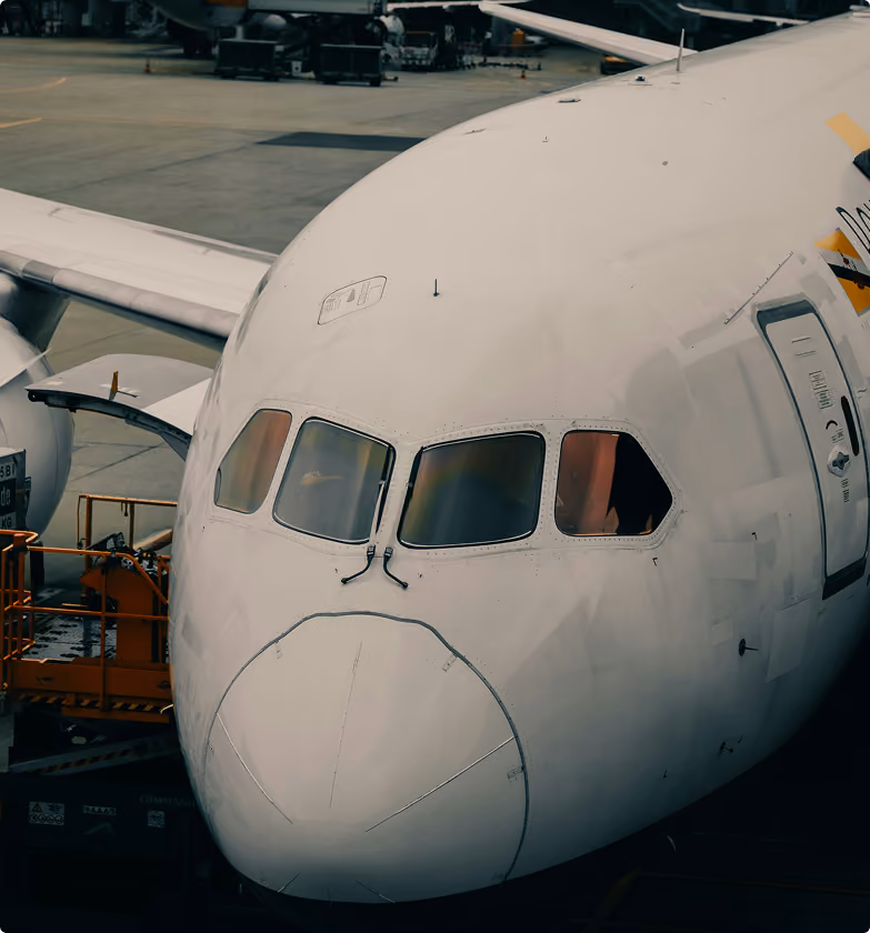 Close-up view of the nose and cockpit windows of a large white airplane parked at an airport gate, with part of the wing and ground equipment visible.